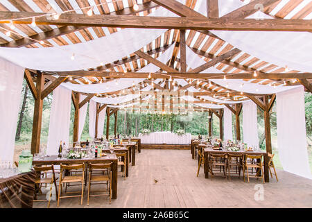 Innenraum einer Hochzeit Tabellen im rustikalen Hütte aus Holz, mit Glühbirnen, bereit für Gäste eingerichtet. Tische im Freien serviert und Veranstaltungsräume mit eingerichtet Stockfoto