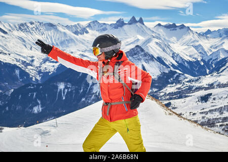 Frau Skifahrer lächelnd und im Les Sybelles Skigebiet posiert, mit Aiguilles d'Arves Gipfeln im Hintergrund, an einem sonnigen Tag mit blauem Himmel. Stockfoto