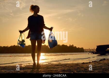 Junge Frau in schwarzem Gummi Handschuhe ist Laufen mit Müllsack entlang einer schmutzigen Ufer des Flusses und Reinigung, Abfall. Sonnenuntergang. Die Menschen und der Ökologie. Ri Stockfoto
