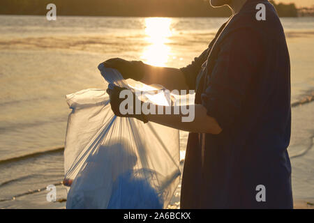 Junge Dame in schwarz Handschuhe ist Laufen mit Müllsack entlang einer schmutzigen Ufer des Flusses und Reinigung, Abfall, alte Glasflaschen. Sonnenuntergang. Menschen und e Stockfoto