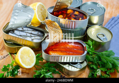 Fisch und Meeresfrüchte, Dosen öffnen Dosen mit sortierten Konserven mit Fisch und Muscheln serviert auf Holztisch mit Petersilie und Pfeffer Stockfoto