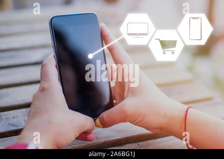 Konzept des Kaufens Elektronik im Internet. Symbole mit Trolley, Smartphone und TV auf dem Hintergrund der Hände halten ein Smartphone. Stockfoto