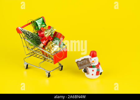 Shopping Cart mit Weihnachtsbaum und Miniatur Geschenkboxen mit Schneemann Puppe auf gelben Hintergrund. Weihnachten und Neujahr Shopping zeit Hintergrund con Stockfoto