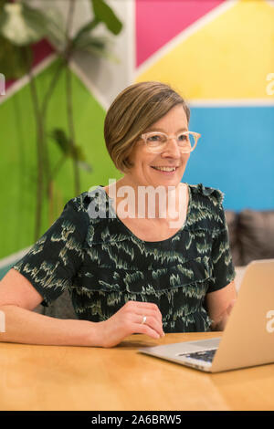 Eine glückliche Frau sitzt an einem Schreibtisch in einem bunten Büro an einem Notebook arbeitet Stockfoto