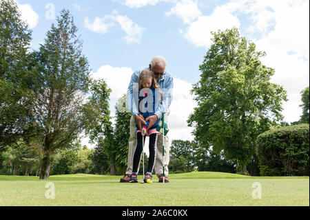 Ein Großvater lehrt seine Enkelin, wie man einen Putter auf einem Golf Putting Green verwendet. Stockfoto