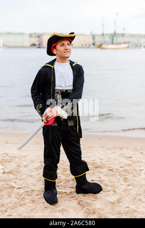 Jungen kaukasischen Mann in Schwarz und Weiß Pirate Kostüm holding Spielzeugschwert und Wegsehen am Sandstrand. Vertikale. Stockfoto