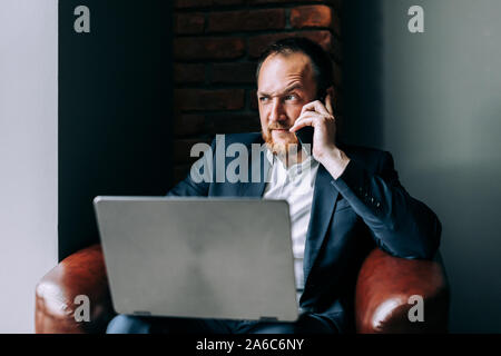 Junge Geschäftsmann im Anzug in einem Stuhl mit einem Laptop sitzen und Telefonieren. Stockfoto