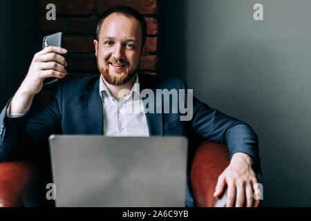 Nach erfolgreicher Geschäftsmann im Anzug in einem Stuhl mit einem Laptop sitzen, halten ein Smartphone in der Hand und lächelt. Stockfoto