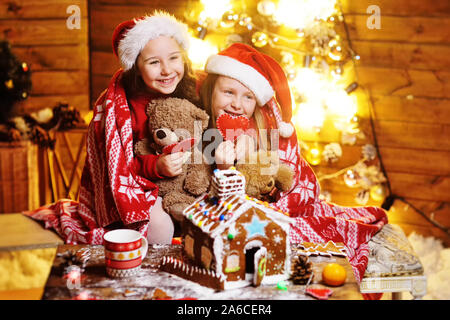 Zwei süße kleine lustige Mädchen in roten Nikolausmützen mit einem plaid Lächeln vor dem Hintergrund von Weihnachten Dekor und Beleuchtung abgedeckt und einen schönen Gin machen Stockfoto