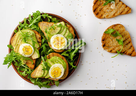 Die richtige Ernährung. Avocado Toast mit Ei und Rucola Salat auf weißem Hintergrund. Ansicht von oben, flach Stockfoto