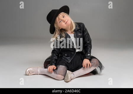 Kleine blonde Mädchen in einem weißen Kleid und schwarzen Lederjacke posiert stehend über einen grauen Hintergrund. Stockfoto