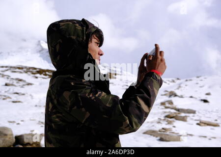 Ein junger Kerl in einem Camouflage Wind Jacke nimmt ein Foto auf einem Smartphone in der schneebedeckten Himalaya Berge an einem sonnigen Tag Stockfoto