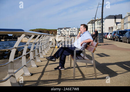 Mittleres Alter Mann sitzt auf einer Bank an der Strandpromenade in Portstewart essen ein Eis an einem sonnigen Herbsttag Stockfoto