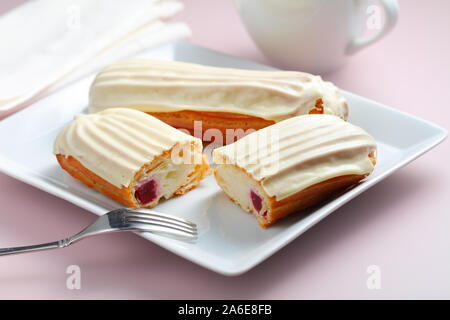 Eclairs mit weißer Schokolade Glasur in eine Schüssel auf rosa Hintergrund. Einer der Kuchen wird halbiert und seinem Querschnitt ist sichtbar Stockfoto