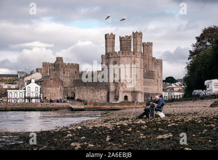 Paar sitzt auf felsigen Strand, Angeln bei Ebbe, mit Caernarfon Castle hinter Stockfoto