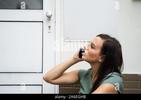 Eine Angst junge Frau sitzt vor der Tür Sprechen auf Mobilephone Stockfoto