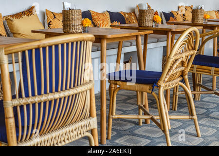 Natürliche Farbe Holz- und Rattanmöbeln auf ein Café im Freien. Blue Kissen, frischen Ringelblume Blumen, strohgedeckten Gewebe Inhaber. Sonnigen morgen. Stockfoto