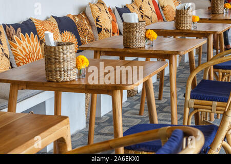 Natürliche Farbe Holz- und Rattanmöbeln auf ein Café im Freien. Blue Kissen, frischen Ringelblume Blumen, strohgedeckten Gewebe Inhaber. Sonnigen morgen. Stockfoto