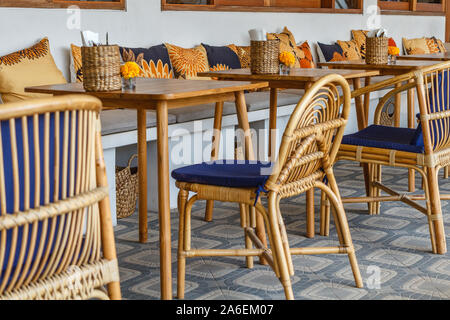 Natürliche Farbe Holz- und Rattanmöbeln auf ein Café im Freien. Blue Kissen, frischen Ringelblume Blumen, strohgedeckten Gewebe Inhaber. Sonnigen morgen. Stockfoto