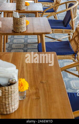 Natürliche Farbe Holz- und Rattanmöbeln auf ein Café im Freien. Blue Kissen, frischen Ringelblume Blumen, strohgedeckten Gewebe Inhaber. Sonnigen morgen. Stockfoto