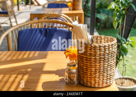 Natürliche Farbe Holz- und Rattanmöbeln auf ein Café im Freien. Blue Kissen, frischen Ringelblume Blumen, strohgedeckten Gewebe Inhaber. Sonnigen morgen. Stockfoto