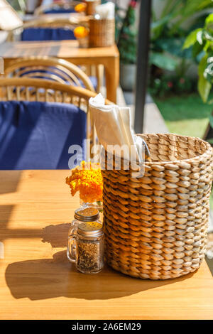 Natürliche Farbe Holz- und Rattanmöbeln auf ein Café im Freien. Blue Kissen, frischen Ringelblume Blumen, strohgedeckten Gewebe Inhaber. Sonnigen morgen. Stockfoto