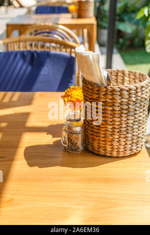 Natürliche Farbe Holz- und Rattanmöbeln auf ein Café im Freien. Blue Kissen, frischen Ringelblume Blumen, strohgedeckten Gewebe Inhaber. Sonnigen morgen. Stockfoto