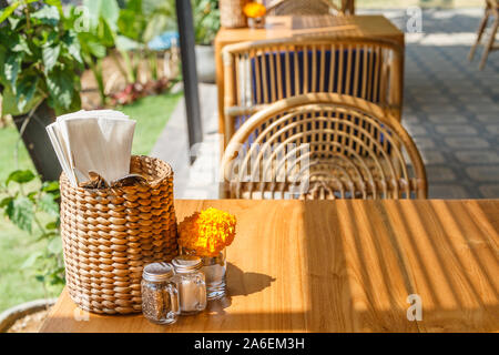 Natürliche Farbe Holz- und Rattanmöbeln auf ein Café im Freien. Blue Kissen, frischen Ringelblume Blumen, strohgedeckten Gewebe Inhaber. Sonnigen morgen. Stockfoto