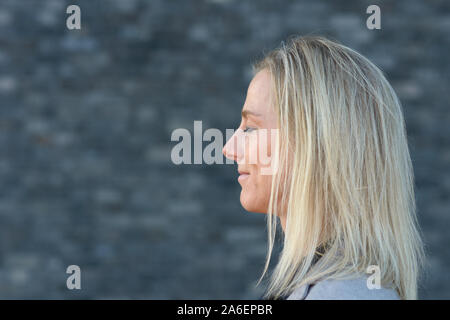 Seite Portrait von geradem Haar blonde Frau in Nahaufnahme, stehen mit geschlossenen Augen und Grinsen auf ihrem Gesicht gegen verwackelte dunkelgrauer Hintergrund mit Stockfoto