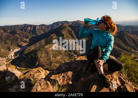 Ein Wanderer Mädchen sitzen und trinken frisches Wasser aus ihren blauen Kantine auf dem Rand der Klippe - Sie trägt eine Sonnenbrille, schwarze Leggings und traine Stockfoto