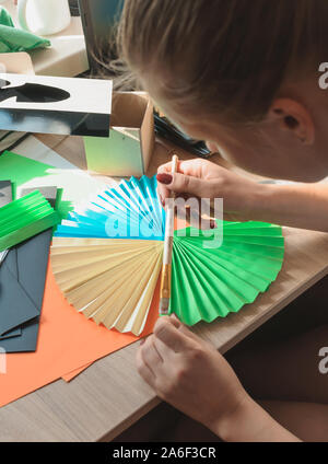 Im Vordergrund weibliche Hände Leim origami Fans, Folien, farbiges Papier, Schere auf einen hölzernen Tisch. Mehrere Leerzeichen für den Ventilator. Für design Vorlage Stockfoto