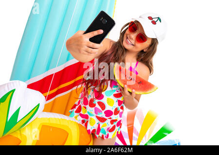Coole Mädchen mit langen gekreuzigt Haar in Sonnenbrille und Sonnencreme, steht in der Nähe von aufblasbaren Matratzen und selfie mit einer Wassermelone tun Stockfoto
