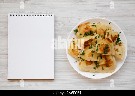 Hausgemachte, traditionelle polnische gebraten Kartoffel pierogis auf einem weissen Teller, leere Notepad, Ansicht von oben. Von oben, flach und Overhead. Kopieren Sie Platz. Stockfoto