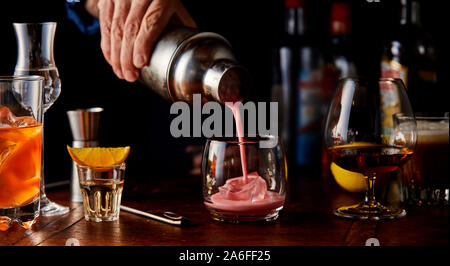 Barkeeper gießen einen fruchtigen Cocktail von einem Shaker auf einer hölzernen Tresen mit einer Auswahl von verschiedenen alkoholischen Getränke in Gläsern Stockfoto