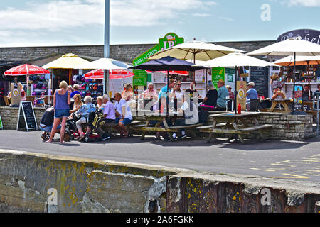 Urlauber und Besucher genießen Sie ein Mittag essen in verschiedenen kleinen Restaurants entlang der Uferstraße an der West Bay. Sonnenschirme bieten angenehmen Schatten. Stockfoto