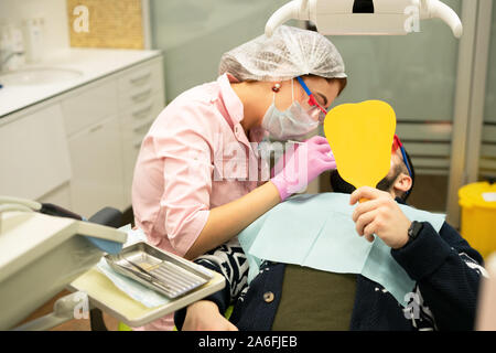 Zahnarzt junge Frau behandelt ein Patient ein Mann. Der Arzt verwendet Einmalhandschuhe, eine Maske, Hut und Schutzbrille. Der Patient schaut in den Spiegel und che Stockfoto