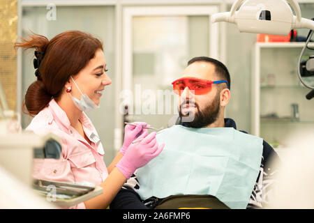 Junge weibliche Zahnarzt, Arzt und Patient Lächeln auf die Kamera an der Zahnklinik. Schutzbrille für den Patienten. Der Arzt in persönliche Schutzausrüstung equi Stockfoto