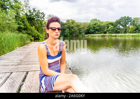 Hübsche junge Modell mit schönen lila Haar sitzt auf einem Holzsteg mit Blick auf den See, die an der Ansicht, gestreifte Kleidung und flache Schuhe Stockfoto