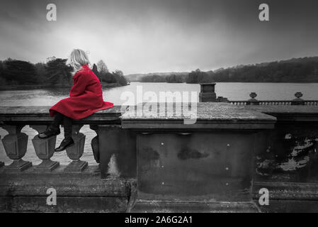 Ein junges Mädchen in einem roten Mantel steht starrte mit Blick auf einen schönen grossen See lehnte sich auf eine Mauer aus Stein, isoliert und allein an Trentham Gardens in Stoke Stockfoto