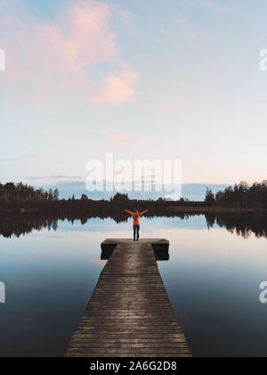 Frau traveler allein stehend auf Pier See Sonnenuntergang und Blick auf den Wald reisen Outdoor Urlaub in Finnland Stockfoto