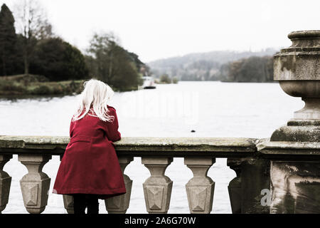 Ein nettes, hübsches kleines Mädchen mit einem roten Mantel mit blonden fließendes Haar blickt, mit Blick auf einen See, allein, in nachdenkliche Isolation, Trentham Gardens Stockfoto