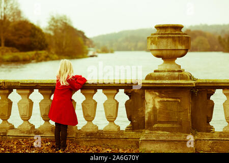 Ein nettes, hübsches kleines Mädchen mit einem roten Mantel mit blonden fließendes Haar blickt, mit Blick auf einen See, allein, in nachdenkliche Isolation, Trentham Gardens Stockfoto