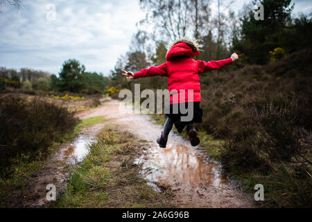 Ein kleines Mädchen geht, läuft und springt durch und in sehr schlammigen Pfützen auf dem Land auf einer Familie zu Fuß trägt einen warmen roten Körper wärmer Stockfoto