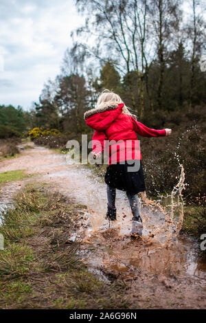 Ein kleines Mädchen geht, läuft und springt durch und in sehr schlammigen Pfützen auf dem Land auf einer Familie zu Fuß trägt einen warmen roten Körper wärmer Stockfoto