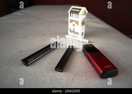 3 vapes Juul, Streben, Breeze, smok infinix mit einer Packung Marlboro Zigaretten und eine Zigarette draußen auf eine weiße strukturierte Tabelle gelegt, isoliert Stockfoto