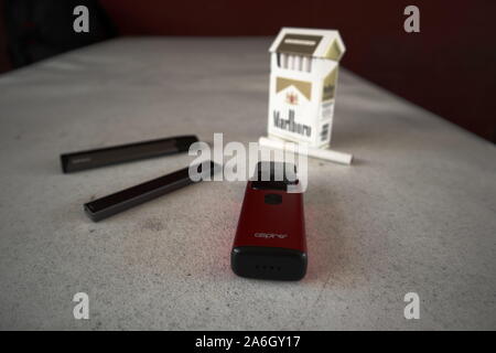 3 vapes Juul, Streben, Breeze, smok infinix mit einer Packung Marlboro Zigaretten und eine Zigarette draußen auf eine weiße strukturierte Tabelle gelegt, isoliert Stockfoto