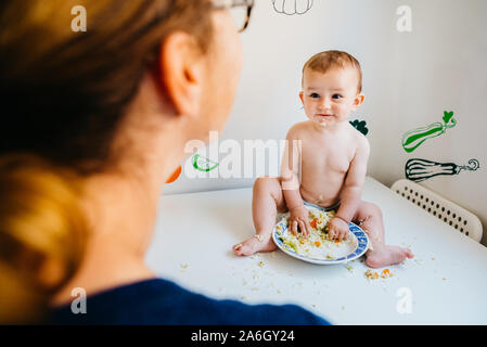 Baby glücklich lächelnd auf seine Mutter sah ihn beim Essen mit den Händen in die Küche. Stockfoto