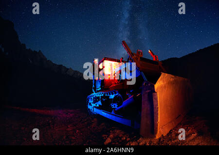 Mann auf Bulldozer mit rot glühenden Kabine und Milchstraße Aussicht nachts Sternenhimmel in den Bergen Stockfoto
