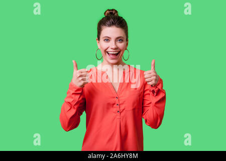 Daumen hoch! Portrait von zufriedenen positive junge Frau mit bun Frisur, große Ohrringe und in rot Bluse ständigen breit lächelnd, wie Geste Stockfoto