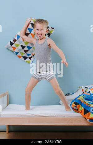 Kleiner Junge stehend auf dem Bett, bereit für eine Kissenschlacht über blaue Wände Stockfoto